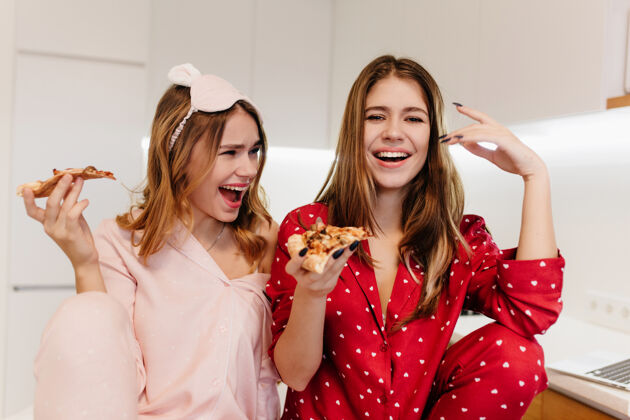 微笑美艳的白种女孩早上在吃早餐时鬼鬼祟祟室内拍摄可爱的姐妹们在吃比萨饼朋友放松卷发