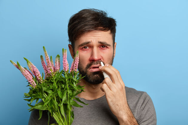 治疗不满意的形象：长胡子的男人眼睛发红 用药水喷鼻涕来治疗打喷嚏和过敏症状 对扳机有反应 眼睛红肿 在室内摆姿势过敏病概念生病敏感男性