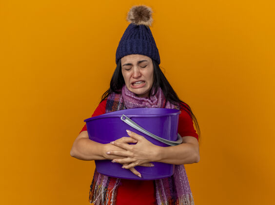 脸恼怒的年轻生病的女人戴着冬天的帽子和围巾拿着塑料桶恶心闭着眼睛隔离在橙色的墙上人抱着人