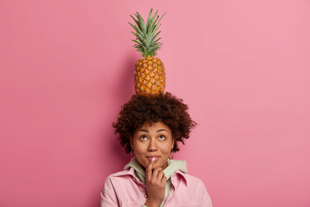 游戏好奇的深色皮肤的非洲裔美国女人头上抱着菠萝 抬头望去 穿着时髦的衣服 对着粉色的墙壁摆姿势 心情积极 玩着异国风味的食物人人类肖像