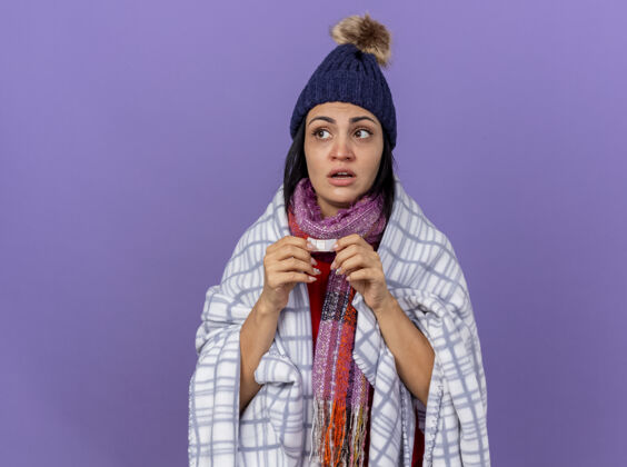 印象印象深刻的年轻生病的女人戴着冬天的帽子和围巾裹着格子布拿着医用石膏看着隔离在紫色墙上的一面年轻人包裹表情