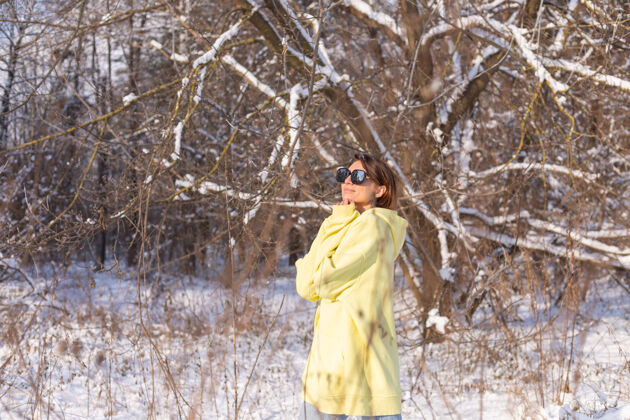 运动阳光明媚的日子里 雪景冬林中一位年轻美女的画像 身着黄色大套头衫 戴着墨镜 享受着阳光和雪时尚自然肖像