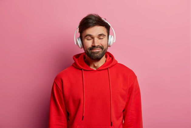 年轻高兴的大胡子男人喜欢用立体声耳机听音乐 闭上眼睛 温柔地微笑 穿着红色运动衫 感觉很好 模特在玫瑰色的粉彩墙上青少年 爱好 生活方式的概念冷静音乐青少年