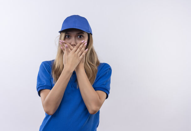 表情害怕的年轻送货女孩穿着蓝色制服 戴着帽子 嘴巴被隔离在白色的手上手手势害怕