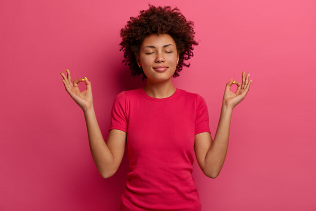 欢呼放松的非洲裔美国妇女的照片显示禅或OK标志 冥想室内 有冷静的表情 闭上眼睛 穿休闲t恤 保持耐心 练习瑜伽练习 在室内摆姿势运动穆德拉冷静
