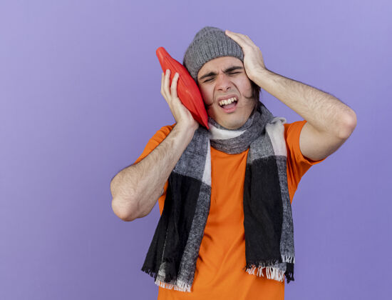 封闭闭着眼睛 戴着带围巾的冬帽的年轻病人 把热水袋放在脸颊上 把手放在疼痛的头上 隔离在紫色的背景上年轻头围巾