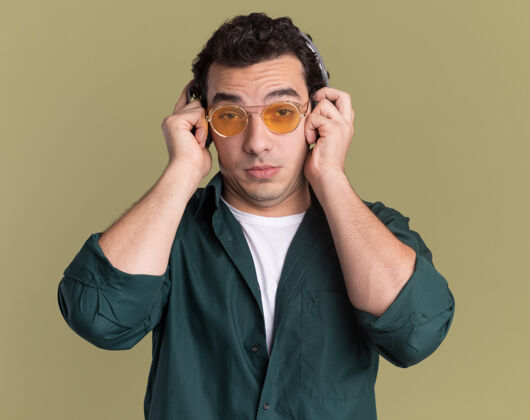 衬衫一个穿着绿衬衫戴着眼镜戴着耳机的年轻人站在绿墙前困惑地看着前方站着年轻戴着