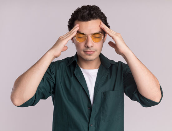 头痛一个穿绿衬衫戴眼镜的年轻人站在白墙上摸着头累得头痛戴着男人疲倦