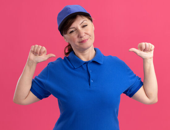 她自己身穿蓝色制服 头戴鸭舌帽的中年女送货员站在粉红色的墙上 看着前面指着自己帽子女人分娩