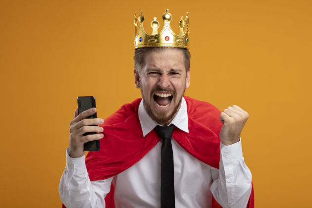 皇冠兴奋的年轻超级英雄戴着领带 戴着王冠 手持手机 在橙色背景上显示“是”的手势是的兴奋超级英雄