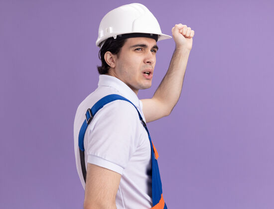 站立年轻的建筑工人穿着建筑制服 戴着安全帽 背着拳头站在紫色的墙上建设者背安全