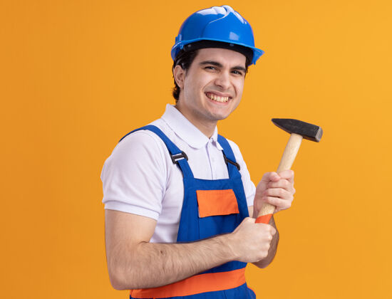 抱着快乐的年轻建筑工人身穿建筑制服 戴着安全帽 手持铁锤 站在橘色的墙上 面带微笑地看着前方欢呼建筑男人