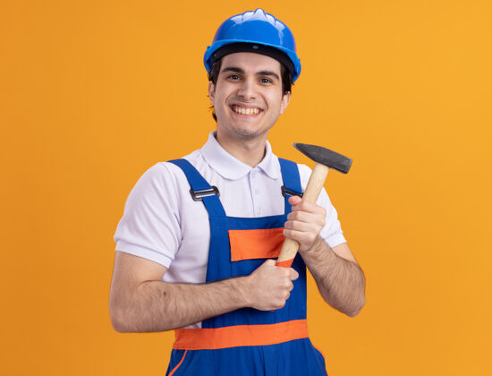 站着快乐的年轻建筑工人身穿建筑制服 戴着安全帽 手持铁锤 站在橘色的墙上 面带微笑地看着前方建筑男人安全