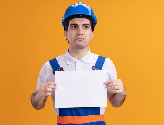 建设者年轻的建筑工人身穿建筑制服 戴着安全帽 拿着空白的一页 站在橙色的墙上 脸上带着悲伤的表情 望向一边空白头盔站在一边