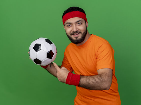 年轻戴着头带和腕带的年轻运动型男子微笑着 指着绿色背景上孤立的球头带运动腕带