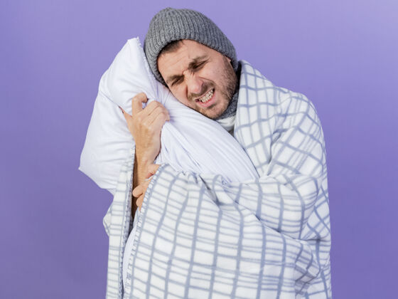格子疼痛的年轻病人戴着冬天的帽子 围巾用格子布包着 抱着紫色背景上孤立的枕头枕头包裹围巾