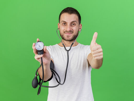 年轻的高兴的年轻病患手持和佩戴听诊器显示拇指孤立在绿色背景拇指请带病拿着