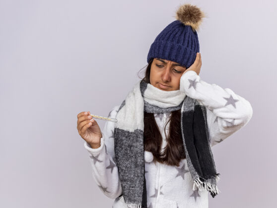 关心带着围巾 戴着冬日帽 拿着体温计 手放在头上 隔离在白色背景上的关心的生病的小女孩年轻人手手