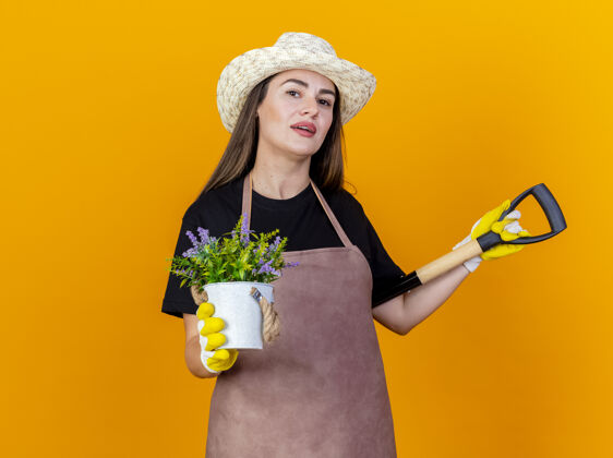 请高兴美丽的园丁女孩穿着制服 戴着园艺手套帽子把铁锹放在肩上 拿着在橙色背景上孤立的花盆里的花帽子手套花园
