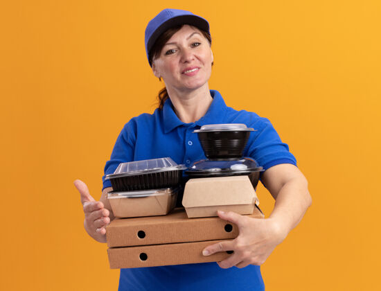 送货快乐的中年送货员身着蓝色制服 戴着帽子 手里拿着披萨盒和食品包 微笑着自信地站在橙色的墙上看着前方拿着女人披萨