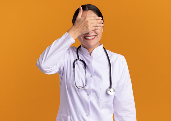 手身穿医用外套的年轻女医生 脖子上戴着听诊器 站在橘色的墙上微笑着闭上眼睛闭着医生女人