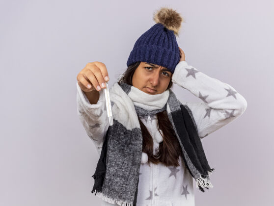 头戴着冬天的帽子 戴着围巾 手放在头上 拿着温度计 站在白色背景上 留着复印空间的相机前 一个未被释放的生病的小女孩疾病年轻帽子