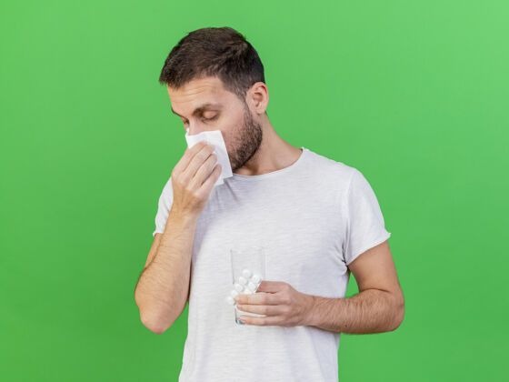 疾病年轻的病人闭着眼睛 用餐巾擦鼻子 拿着药片 一杯水隔离在绿色的背景上药片拿着湿巾
