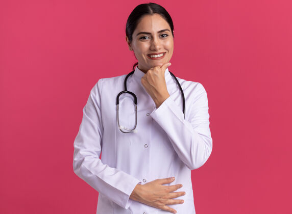外套年轻的女医生穿着医用外套 手持听诊器 站在粉红色的墙上 微笑着自信地看着前方微笑医生医生