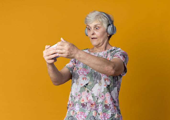 拿着惊讶的老妇人拿着耳机 看着电话孤零零地挂在橘黄色的墙上惊喜老人耳机