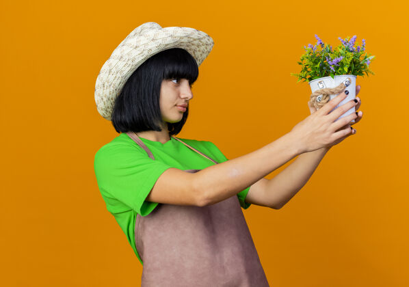 女性自信的年轻女园丁穿着制服 戴着园艺帽 手持花盆 看着花盆花园橙色帽子