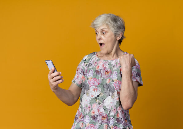 女人惊讶的老妇人举起拳头看着隔离在橙色墙上的电话老的提高电话