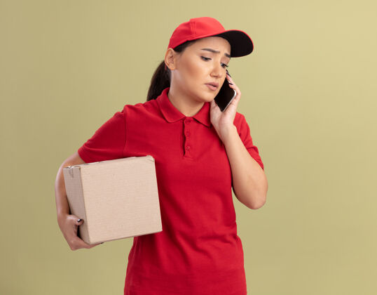 帽子站在绿色的墙上 一位身穿红色制服 戴着帽子 拿着纸板箱的年轻女送货员正在讲手机 看上去很困惑女人拿着电话