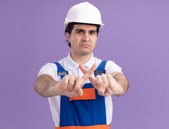 年轻年轻的建筑工人身穿建筑制服 头戴安全帽 表情严肃 停止手势 食指交叉 站在紫色的墙上停车制服严肃