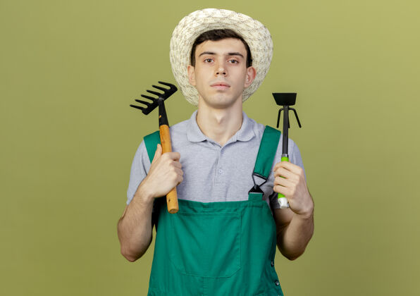 举行自信的年轻男园丁戴着园艺帽拿着耙子和锄头耙子园艺耙花园