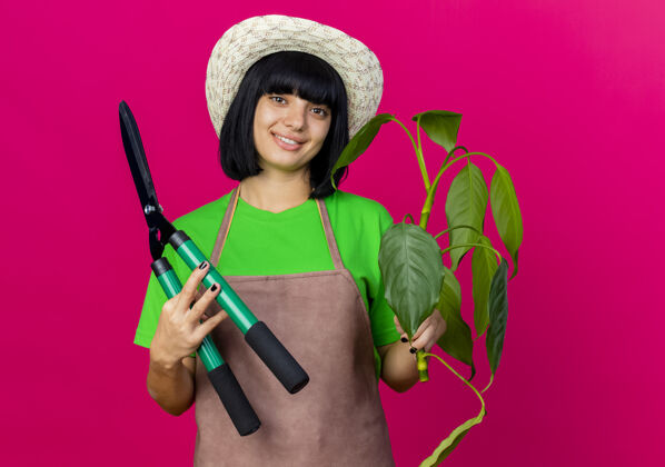 年轻微笑着的年轻女园丁穿着制服戴着园艺帽拿着剪子和植物园艺制服女