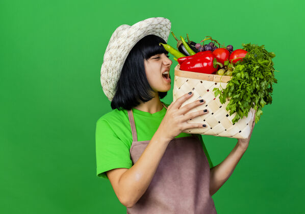 园艺愤怒的穿着制服的年轻女园丁戴着园艺帽 手里拿着一个菜篮子篮子绿色愤怒