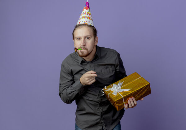 点惊讶的帅哥戴着生日帽 指着礼盒吹着紫色墙上孤立的哨子盒子紫色生日