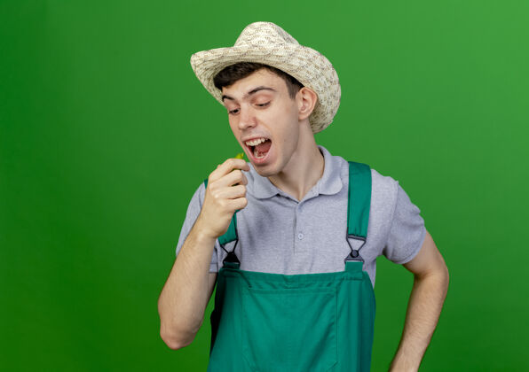 帽子有趣的年轻男园丁戴着园艺帽 看起来和假装咬辣椒隔离在绿色背景与复制空间咬辣园艺