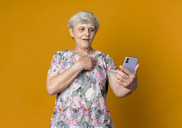 手惊讶的老妇人把手放在胸前看着隔离在橙色墙上的电话老人女人电话