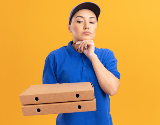 思考身穿蓝色制服 戴着帽子的年轻送货员拿着比萨饼盒 脸上带着沉思的表情 站在橙色的墙上思考着递送制服帽子