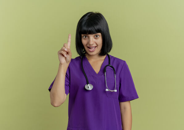 黑发惊讶的年轻黑发女医生穿着制服用听诊器指着了制服橄榄女