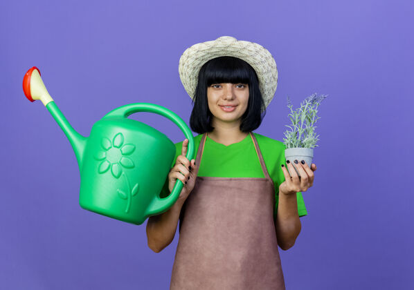 复制身着制服的年轻女园丁微笑着 戴着园艺帽 手里拿着花盆和浇水罐举行制服帽子