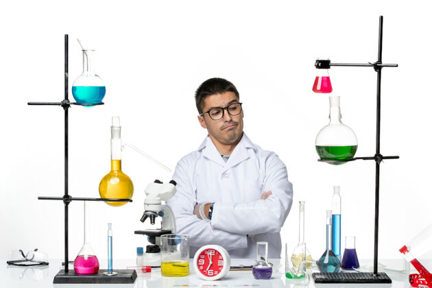 坐姿前视图穿着白色医疗服的男性化学家坐在白色背景病毒科学冠状病毒-大流行实验室准备工作正面科学烧杯