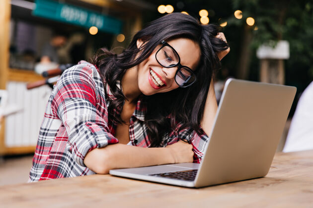 青年戴着眼镜的时髦女孩在使用笔记本电脑时表达积极的情绪可爱的黑发学生做作业的户外肖像可爱乐趣女性