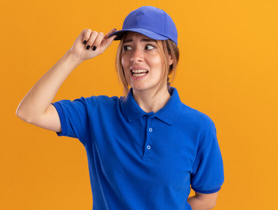 漂亮焦急的年轻漂亮的女送货员穿着制服 把手放在帽子上 看着橙色的一面手焦虑制服
