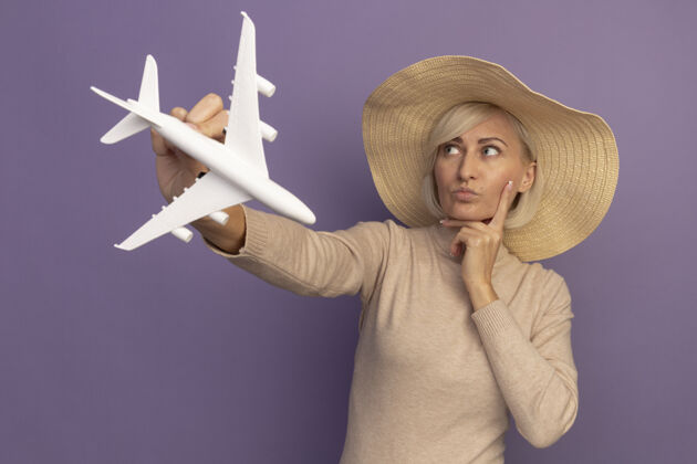 壁板体贴漂亮的金发斯拉夫女人戴着沙滩帽把手放在下巴上拿着模型飞机看着紫色的一面金发飞机年轻