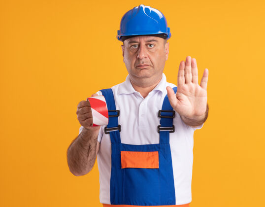 制服自信的白人成年建筑工人穿着制服 拿着透明胶带 在橙色上做着手势停止手势手势磁带手