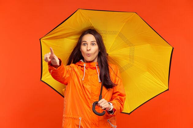 单独这个快乐微笑的年轻女孩穿着秋天的橘色夹克在摄影棚摆姿势 指着左边的红色隔离人类积极的情感寒冷天气的概念女性时尚的概念情绪外套季节