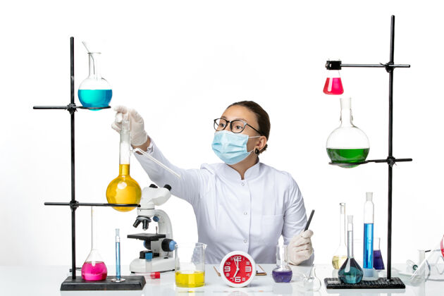 专业前视图穿着白色医疗服的女化学家戴着口罩正坐在白色办公桌上化学家病毒冠状病毒-飞溅实验室套装医学医学