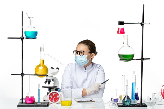 笔记前视图穿着医疗服的女化学家戴着面罩坐在一个白色背景上写笔记飞溅实验室病毒化学冠状病毒写作面具西装
